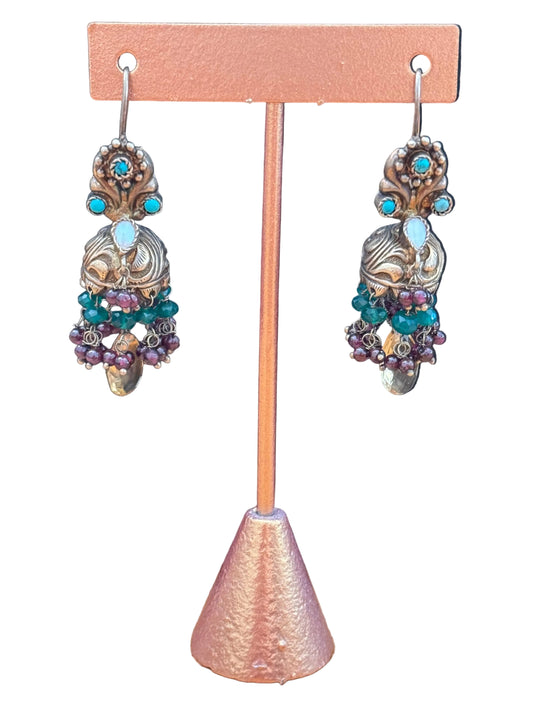 A pair of ornate dangling earrings displayed on a small stand. The earrings have a gold-toned filigree base with turquoise blue stones, a light blue oval stone, and clusters of green and purple beads, set against a black background with a wood surface on the right.